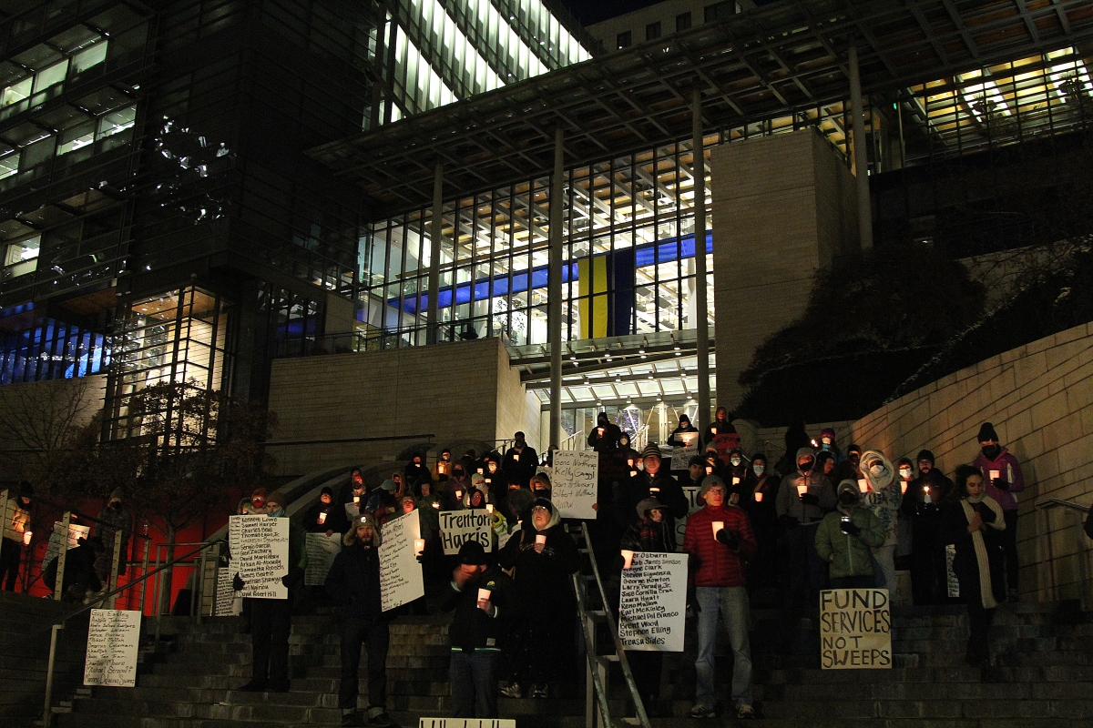 Large group of people standing on steps of lit-up city building at night, holding signs and candles