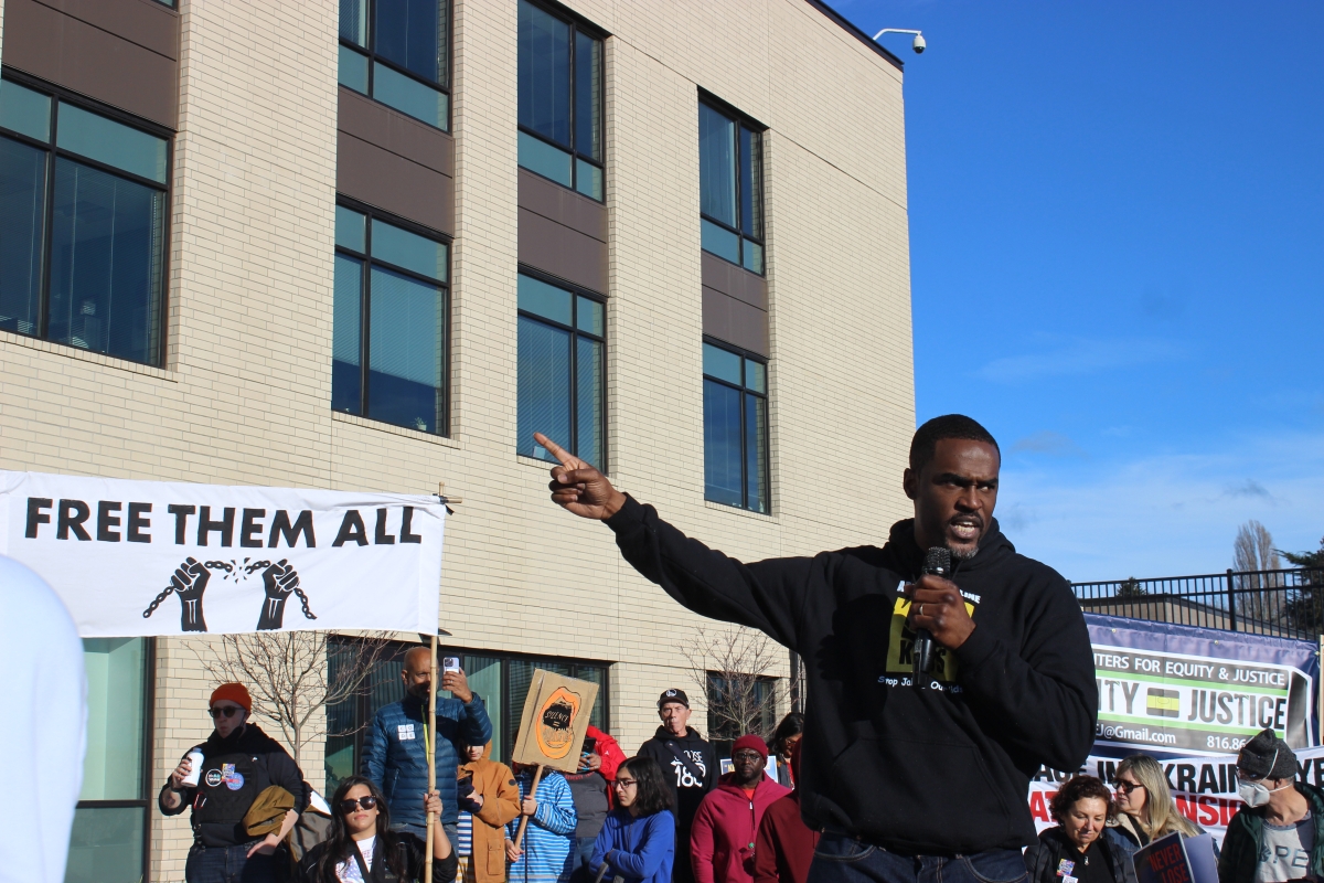 A young/middle-aged Black man in black sweat jacket stands among crowds with signs outside youth jail, speaking into a mic.