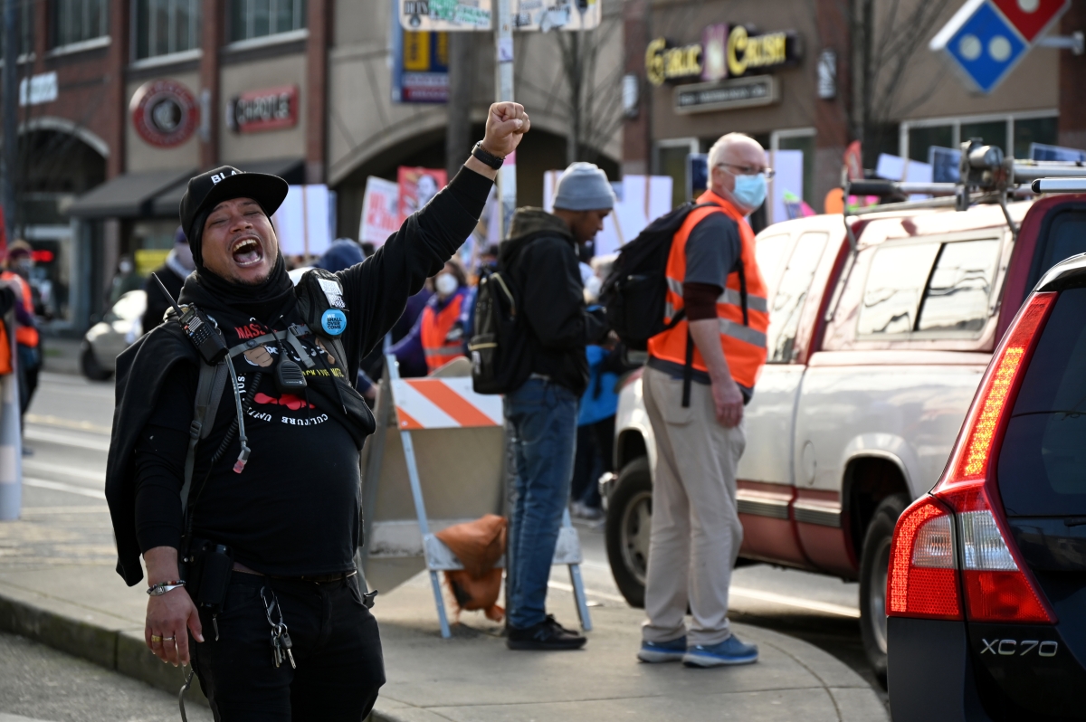 Black man wearing black cap, jacket, walkie-talkie, shouts and holds fist in air on a city street, with other marchers and signs visible behind him.