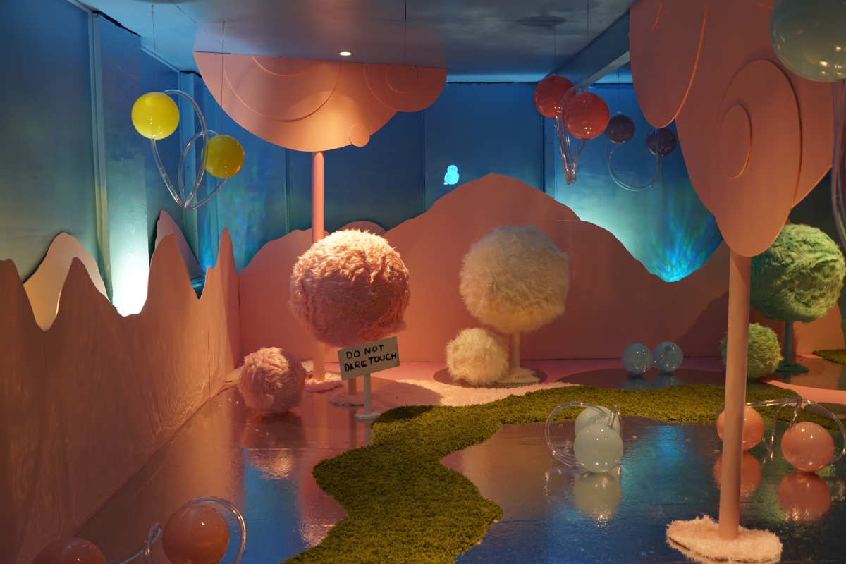 Installation in hues of orange, yellow, green, and blue, showing glass balls on floor in shallow puddles of water as well as furry-looking balls on sticks, with a sign, "Do Not Dare Touch."