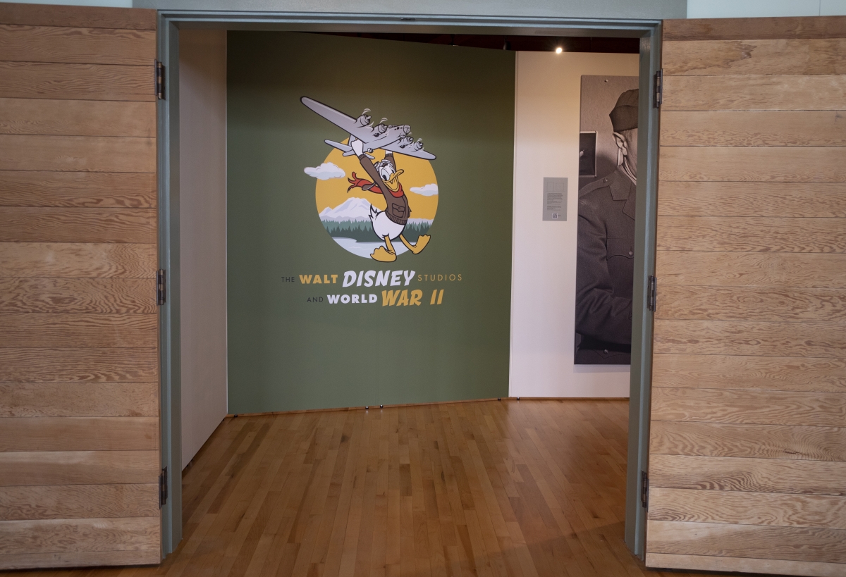 Photograph of entrance panel to exhibit about Walt Disney and World War II, showing Donald Duck hanging from the body of a fighter plane
