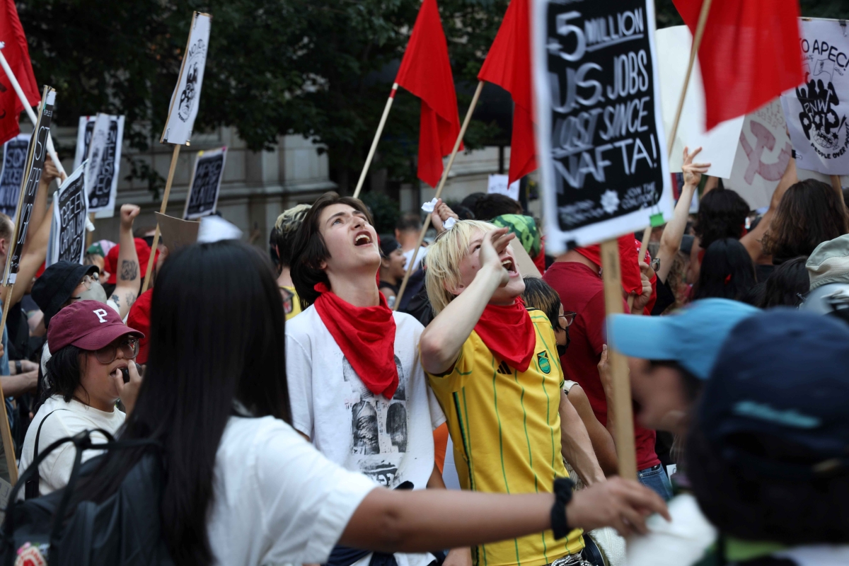 Two young people in red neck bandannas stand surrounded by a crowd holding red flags and protest signs, heads back and mouths open.