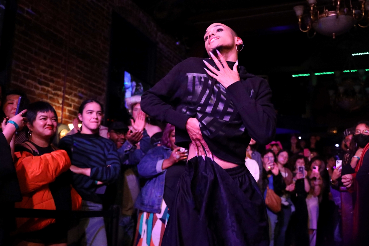 A young, brown-skinned drag queen, seen from below, wearing hoop earrings, black sweatshirt, and long pointy fingernails strikes a pose with hand to chin.