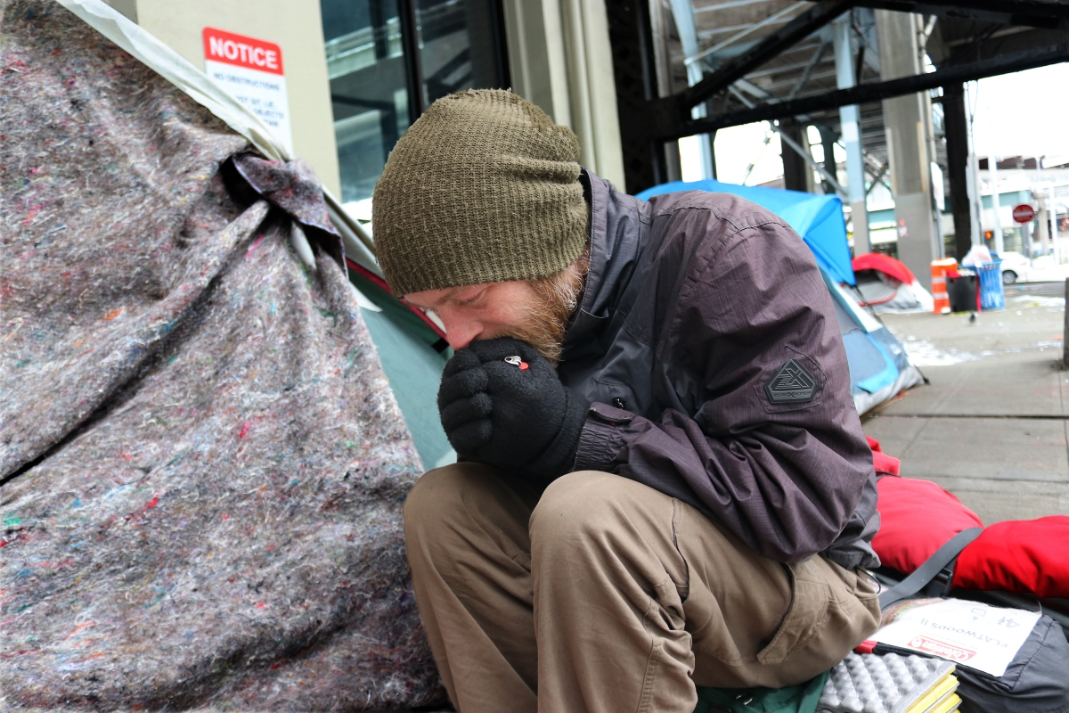 A man wearing a beanie and gloves and sitting next to a tent on a sidewalk rubs his hands together and blows on them during a cold weather encampment sweep.