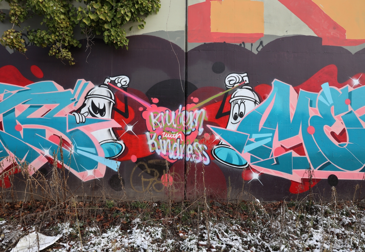 Wall covered with graffiti in shades of red and blue, featuring two spray-paint can characters facing each other while spraying a message in the center reading, "Kill 'Em with Kindness."