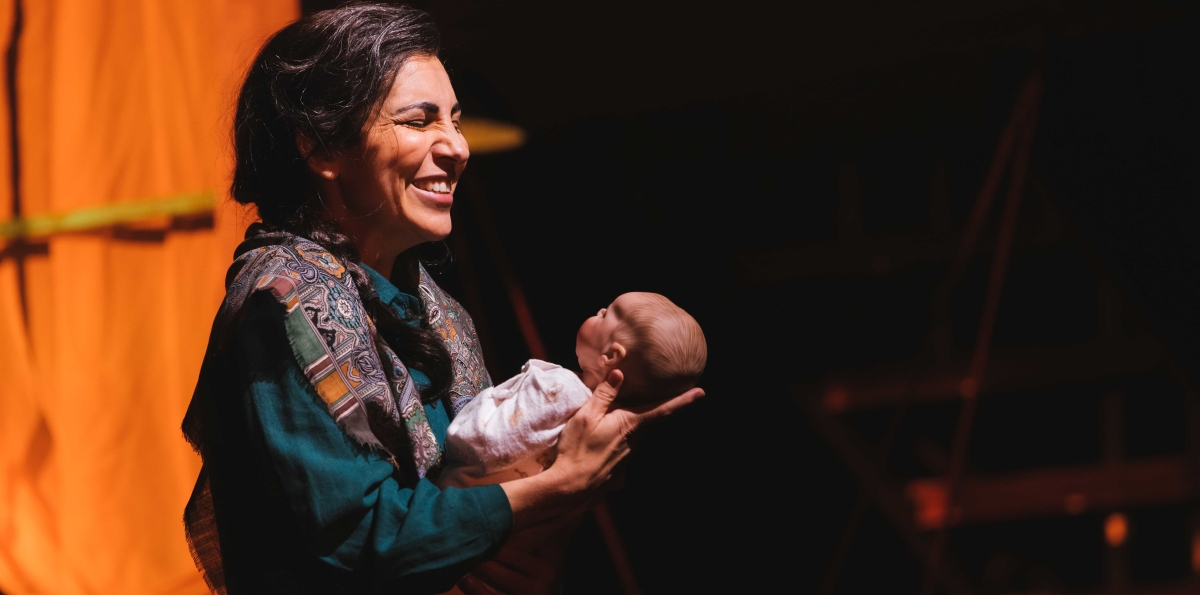 On stage, a woman with long dark hair, in blue dress with print shawl, smiles widely while holding a baby.
