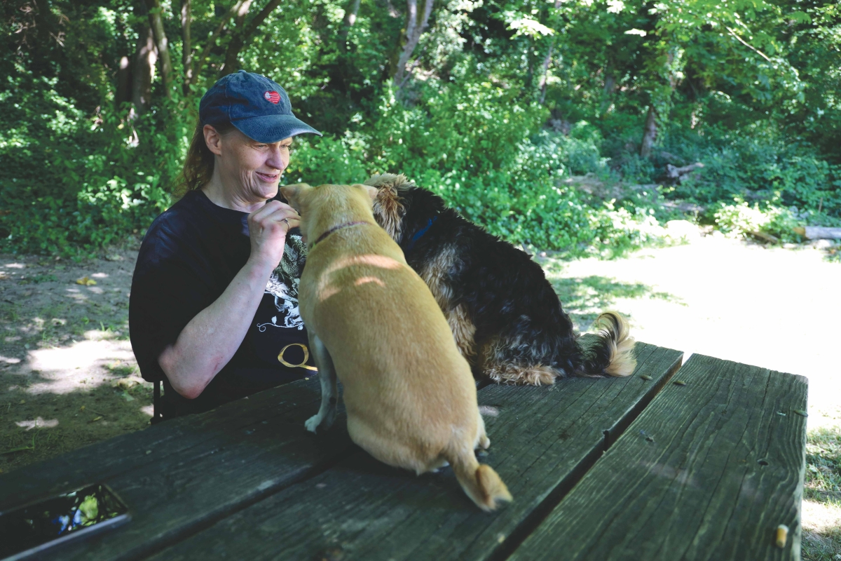 A woman in a ballcap sits at a picnic table, while playing with two small dogs that are sitting on the table; one is tan with short hair and the other is black and tan with fluffier terrier hair.