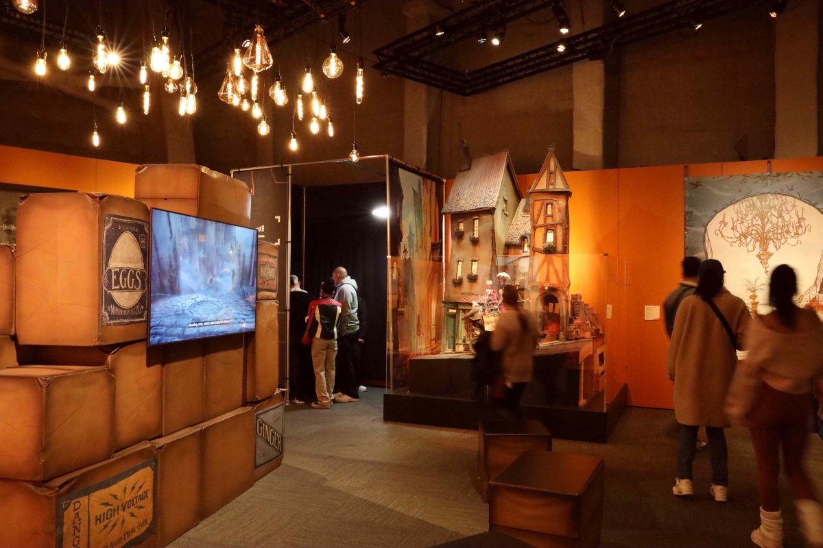 People stand in amber-lit gallery space looking at exhibits including a mockup of tall Tudor-style buildings and a stack of old-fashioned looking warehouse boxes.