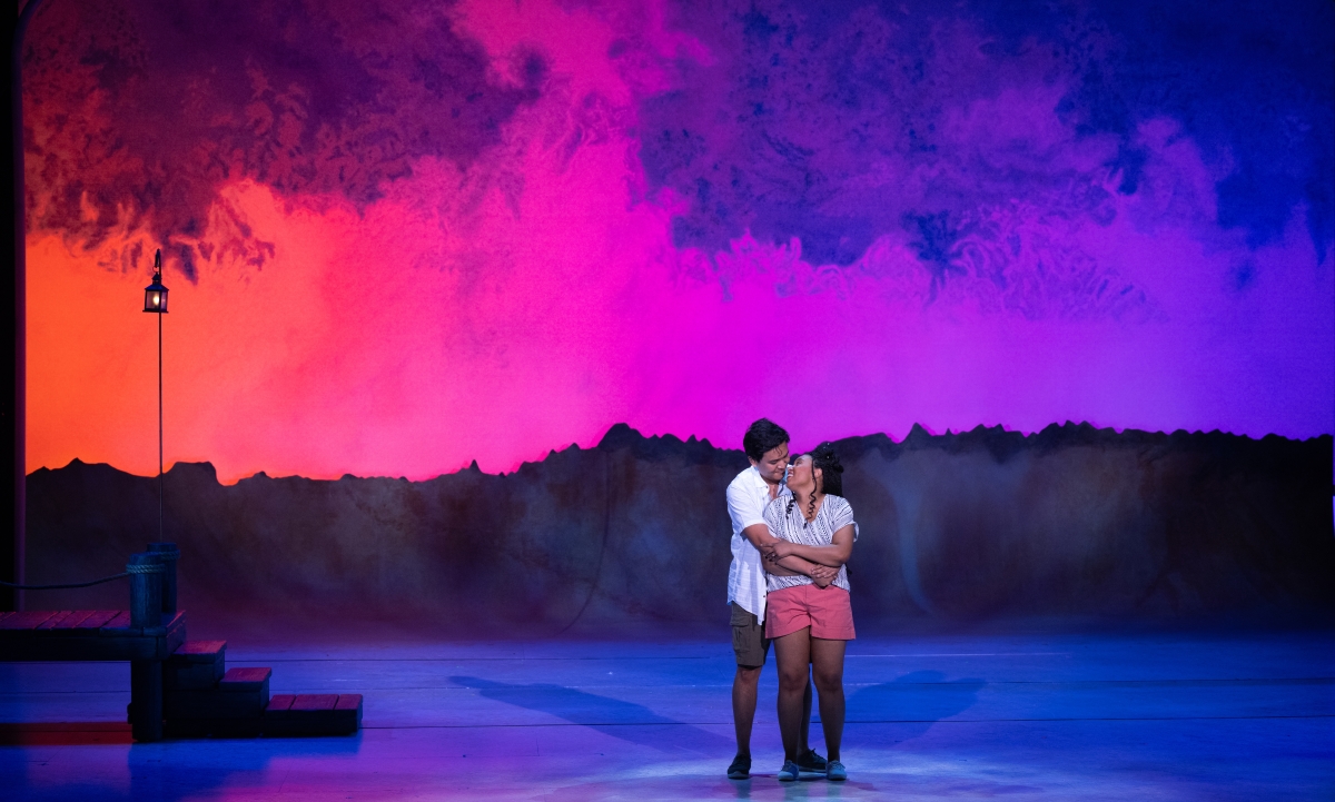 The two lead actors from Village Theatre's "Mamma Mia!" directed by Faith Bennett Russell embrace on stage, in front of a purple background with silhouetted mountains, wearing shorts and light summer button downs.