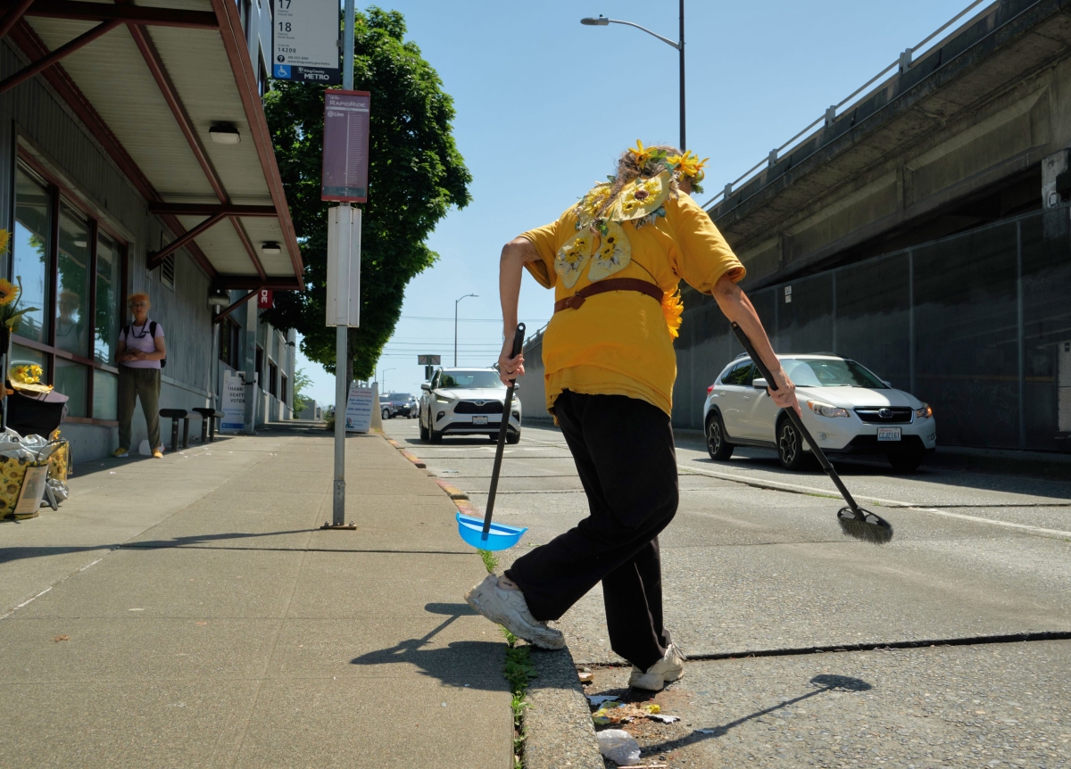 Photograph of woman, seen from behind, wearing yellow shirt and crown of yellow flowers, holding broom and dustpan while stepping off a city street curb on a sunny day.
