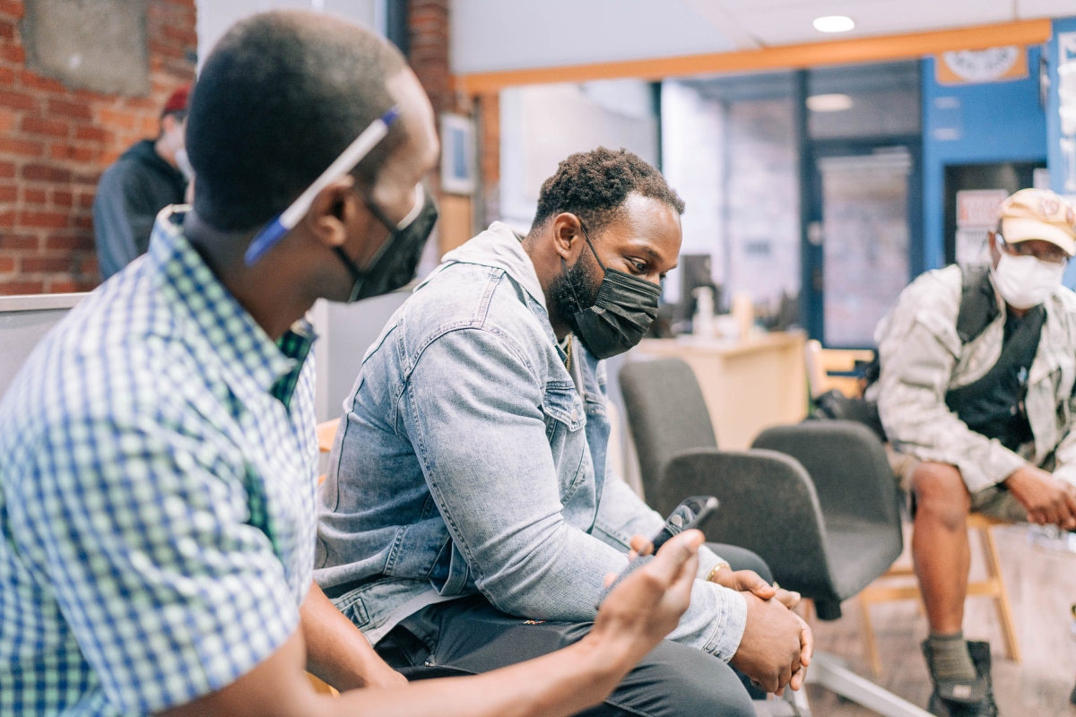 Editor Marcus Harrison Green, holding a phone/recorder and with pen behind his ear, sits next to ex-Seahawk Robert Turbin, who is wearing a denim jacket and black face mask and looking pensive. looking  