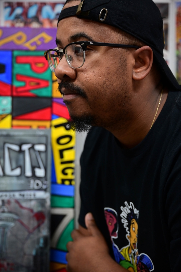 Painter Rodney King, young Black man wearing glasses and backwards baseball cap, photographed in profile in front of paintings