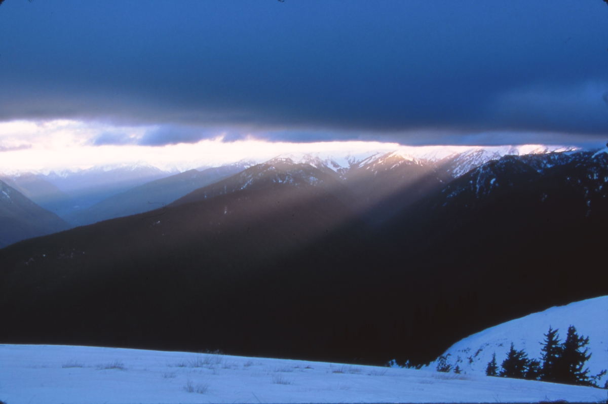 Snowy mountain landscape with hills in foreground, low light over mountains in background