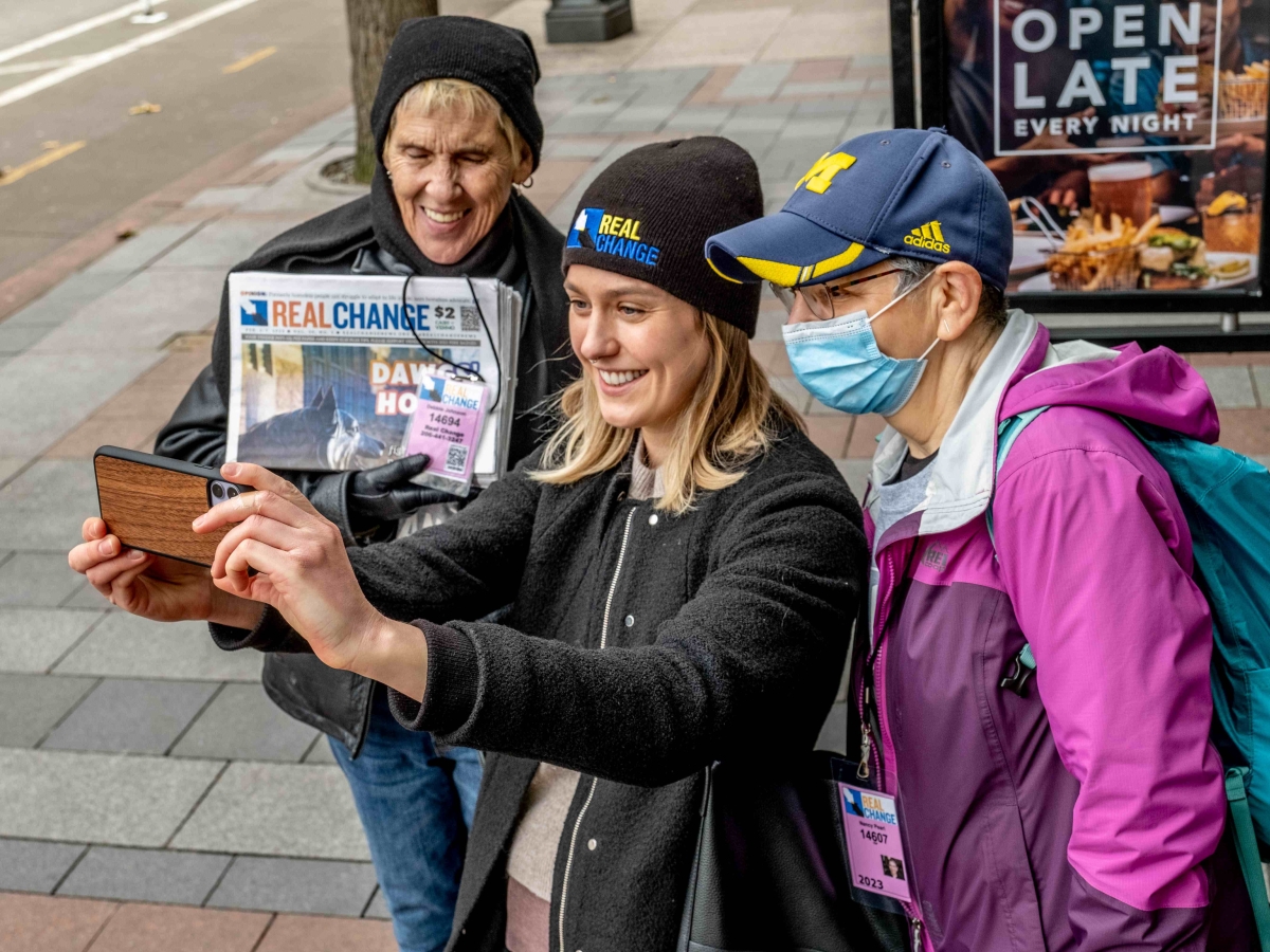 Three women in hats and jackets, one holding Real Change newspapers and one holding up a phone, pose for a selfie.