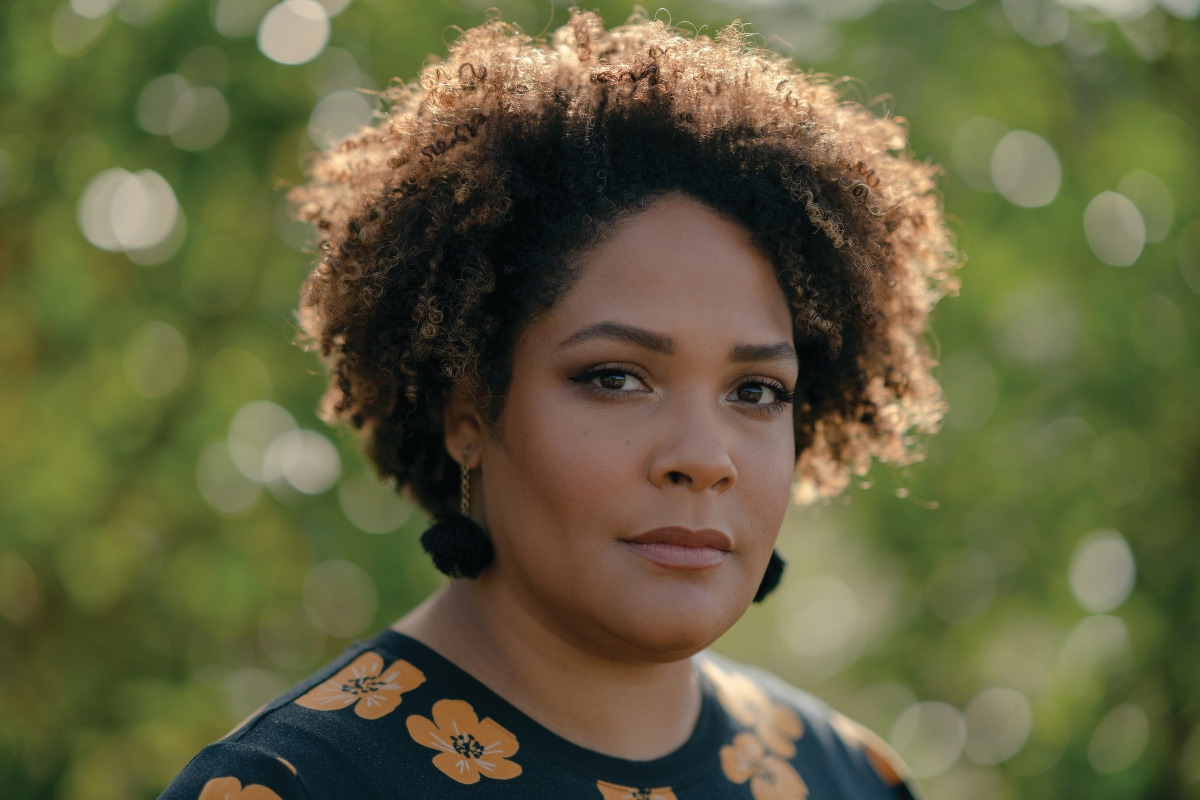 A woman looks proudly into the camera, with her natural hair backlit by golden light. She wears tassel earrings and a blue floral shirt and stands before greenery.