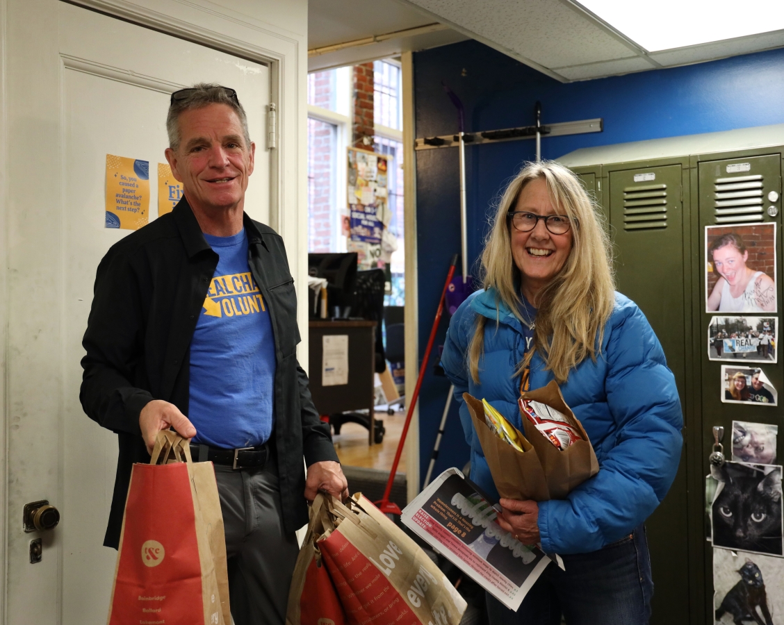 Man with sunglasses on top of head and dressed in jacket and blue Real Change shirt stands next to woman in glasses, with long blonde hair, dressed in blue puffy jacket, both holding paper bags, next to bank of lockers.