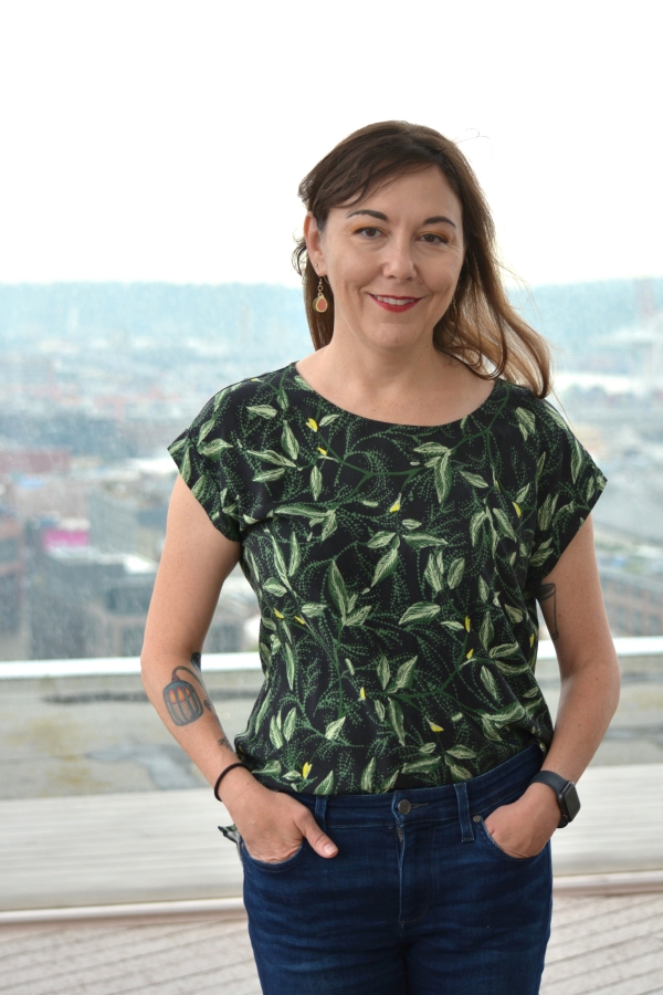 Erica Barnett, young to middle-aged white woman with shoulder-length brown hair and brown eyes, wearing green leaf-print top, poses against view of landscape