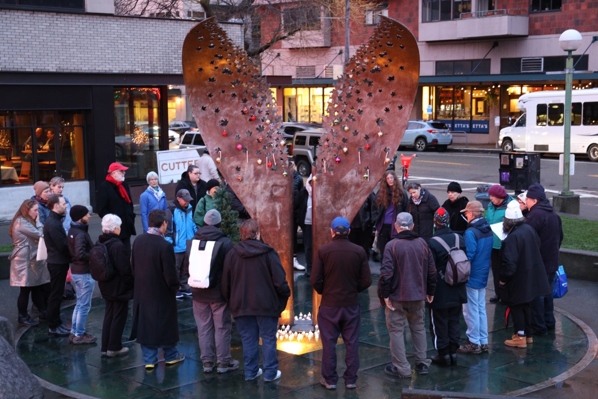 A large group of people in hats and coats stand around a pink sculpture in the shape of two halves of a broken heart, in an outdoor urban location.