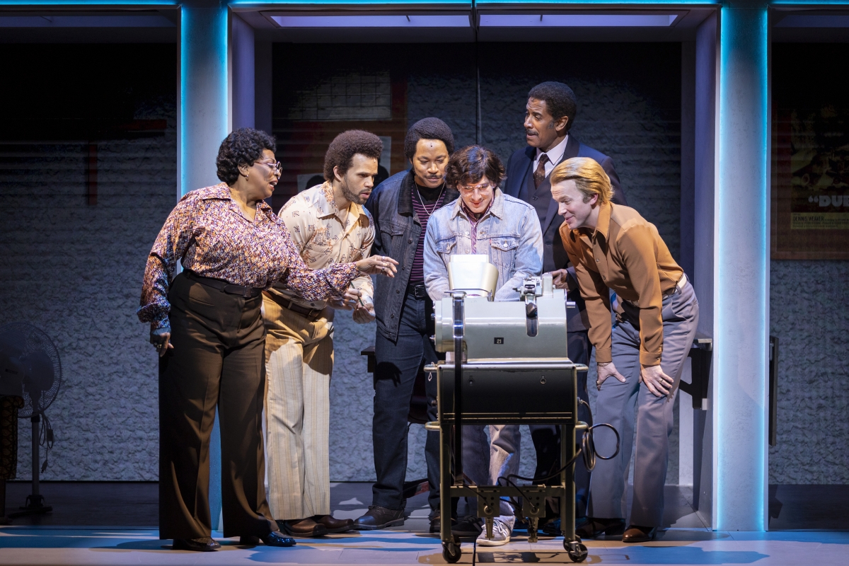 Six people on a stage set gather around a boxy-looking implement on a table.