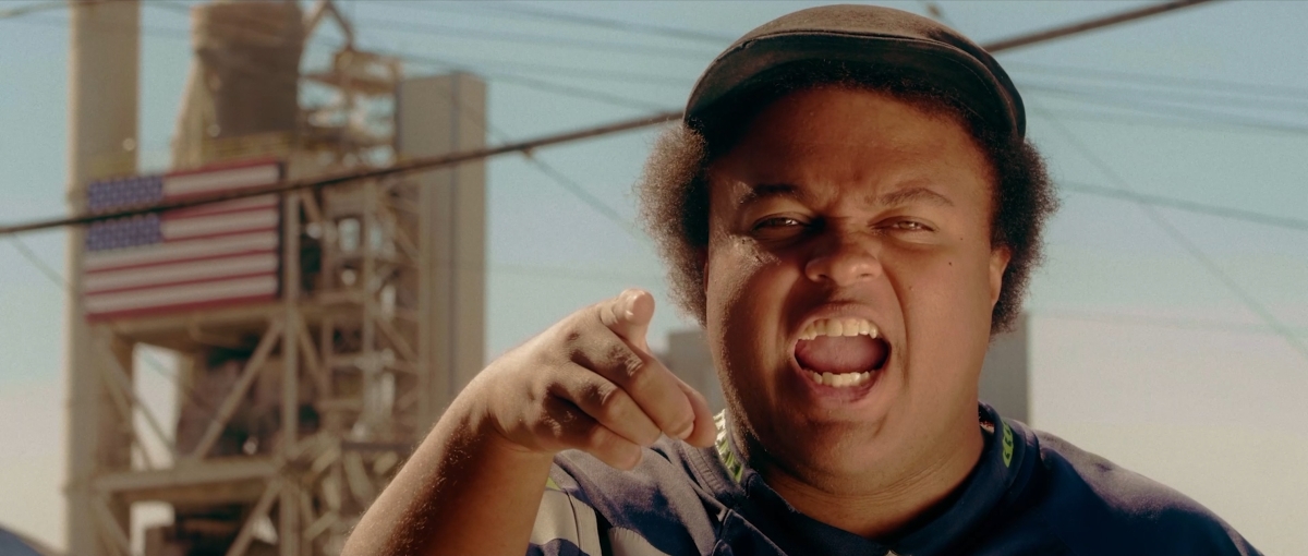 Rapper Fantasy A, a stocky Black man, points at the camera while wearing a Seahawks jersey and a newsboy cap with his fuzzy hair sticking out of the sides