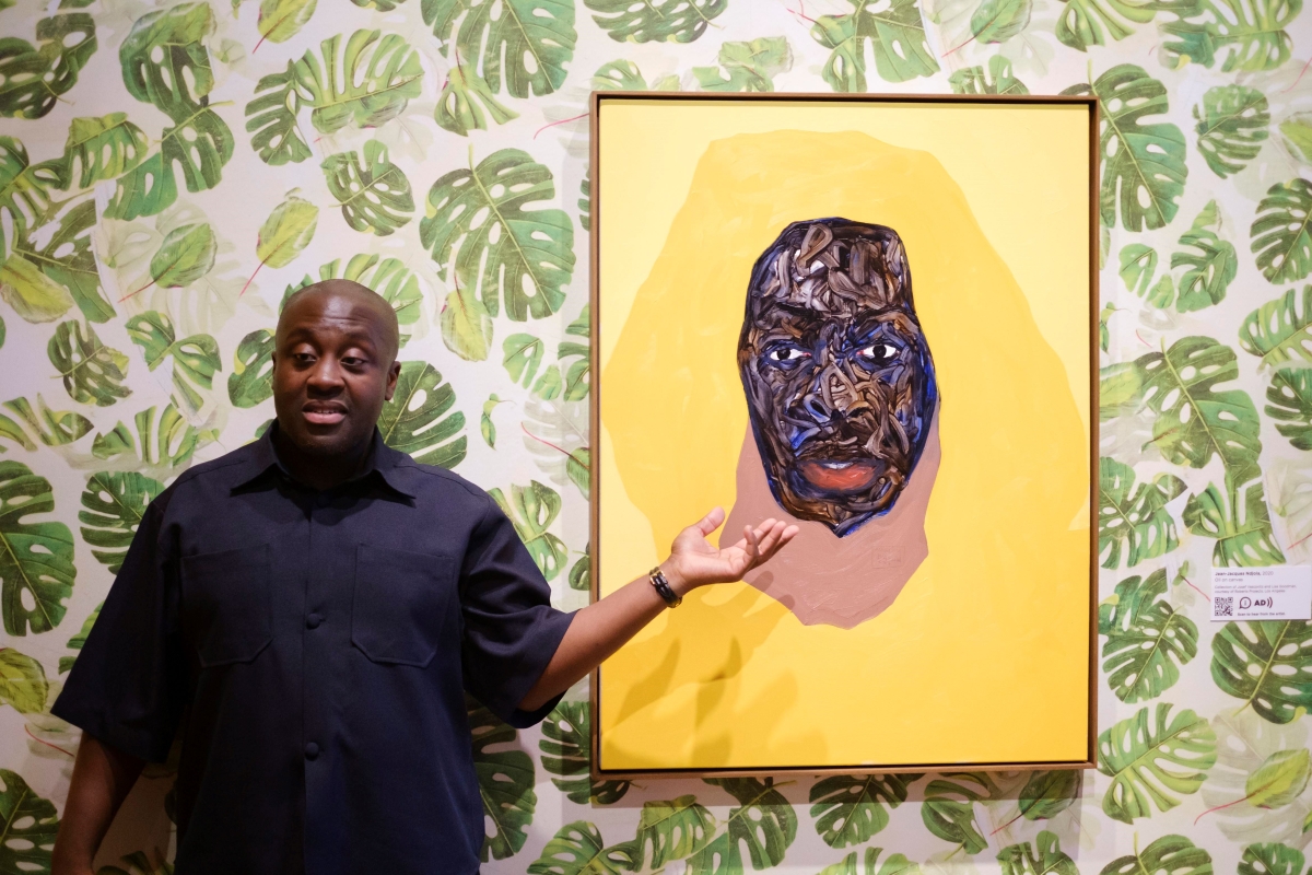 Black man with shaved head, wearing dark short-sleeved shirt, stands next to painting of a Black man's face