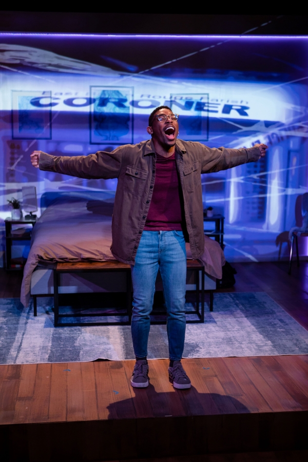 Young Black man wearing glasses and barn jacket stands on stage set, with bedroom furniture in background, with broad smile and holding arms wide 