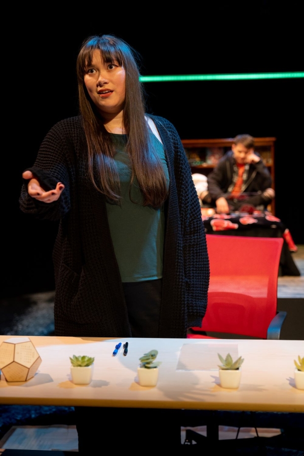 Young white woman with long dark hair, dressed in T-shirt and baggy sweater, stands behind a table lined with succulent pots, holding out her hand.