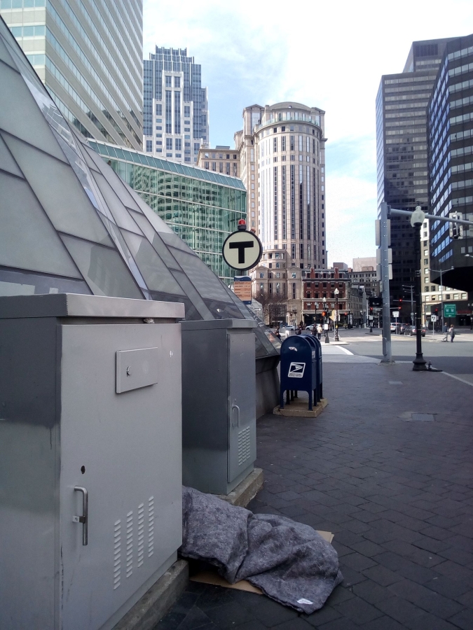 Photograph of exterior of a "T" transit station on a plaza in Boston. A blanket and cardboard flat are visible in foreground.