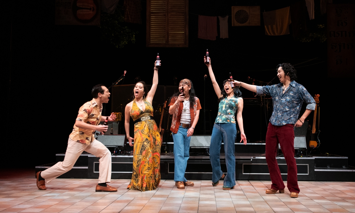 Five people dressed in hippie-ish/psychedelic-print garb hold up bottles and strike celebratory poses on a stage set.