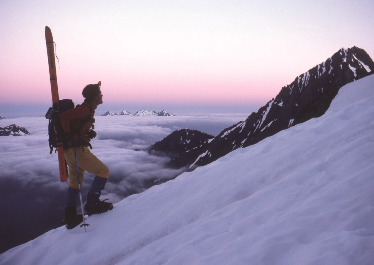 Young white man wearing pack and heavy boots, seen in profile on snowy hill in mountains at sunrise