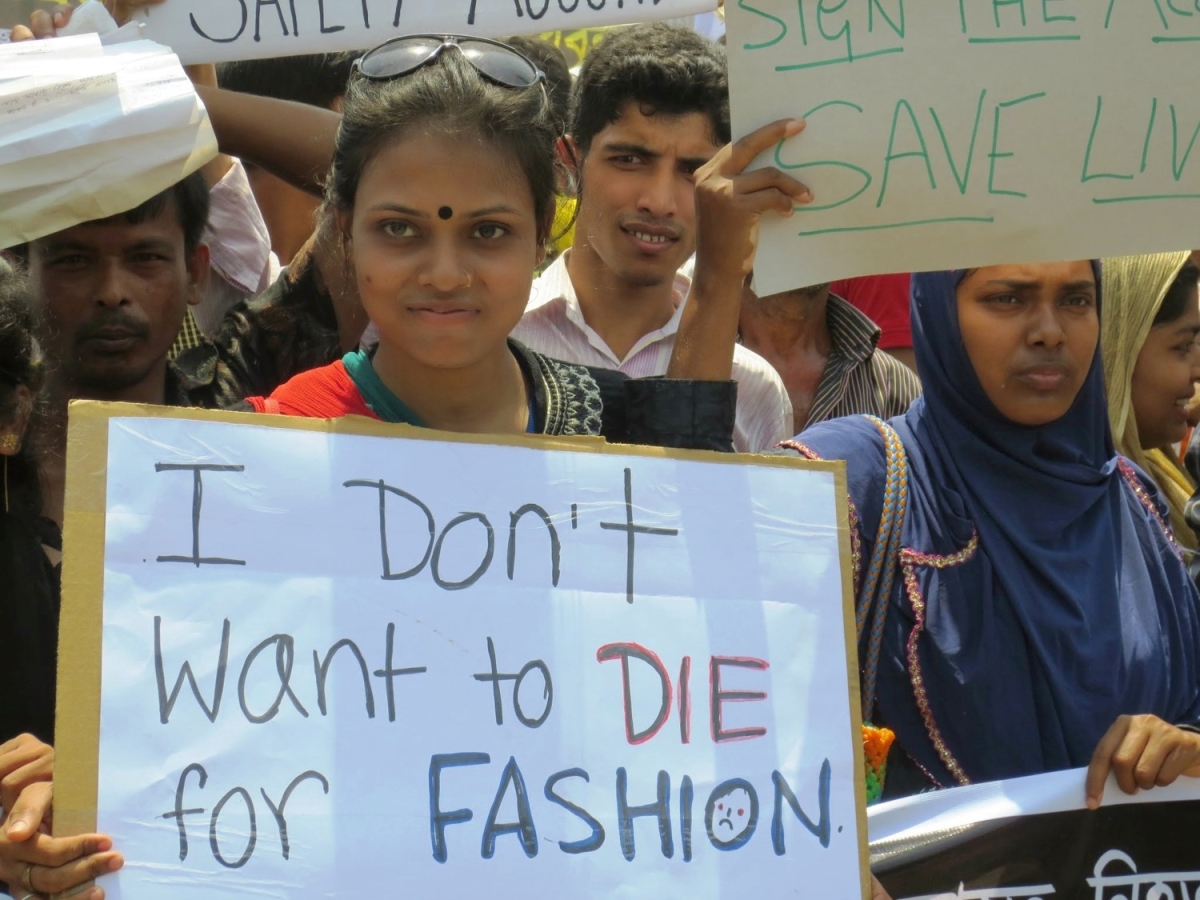 Young South Asian woman backed by others at a rally, holding a sign that reads, "I Don't Want to Die for Fashion."