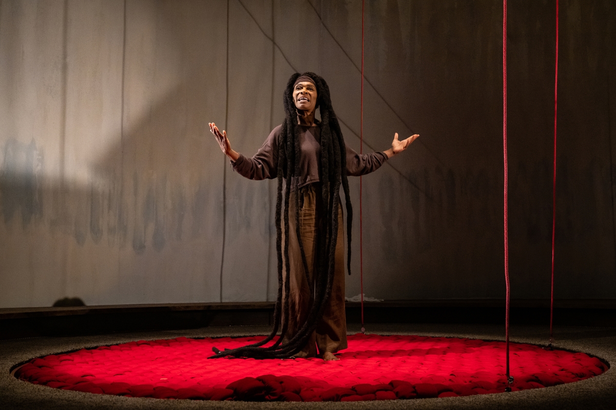 Black woman with floor-length dreadlocks and wearing long dark tunic stands in center of stage with hands outspread.