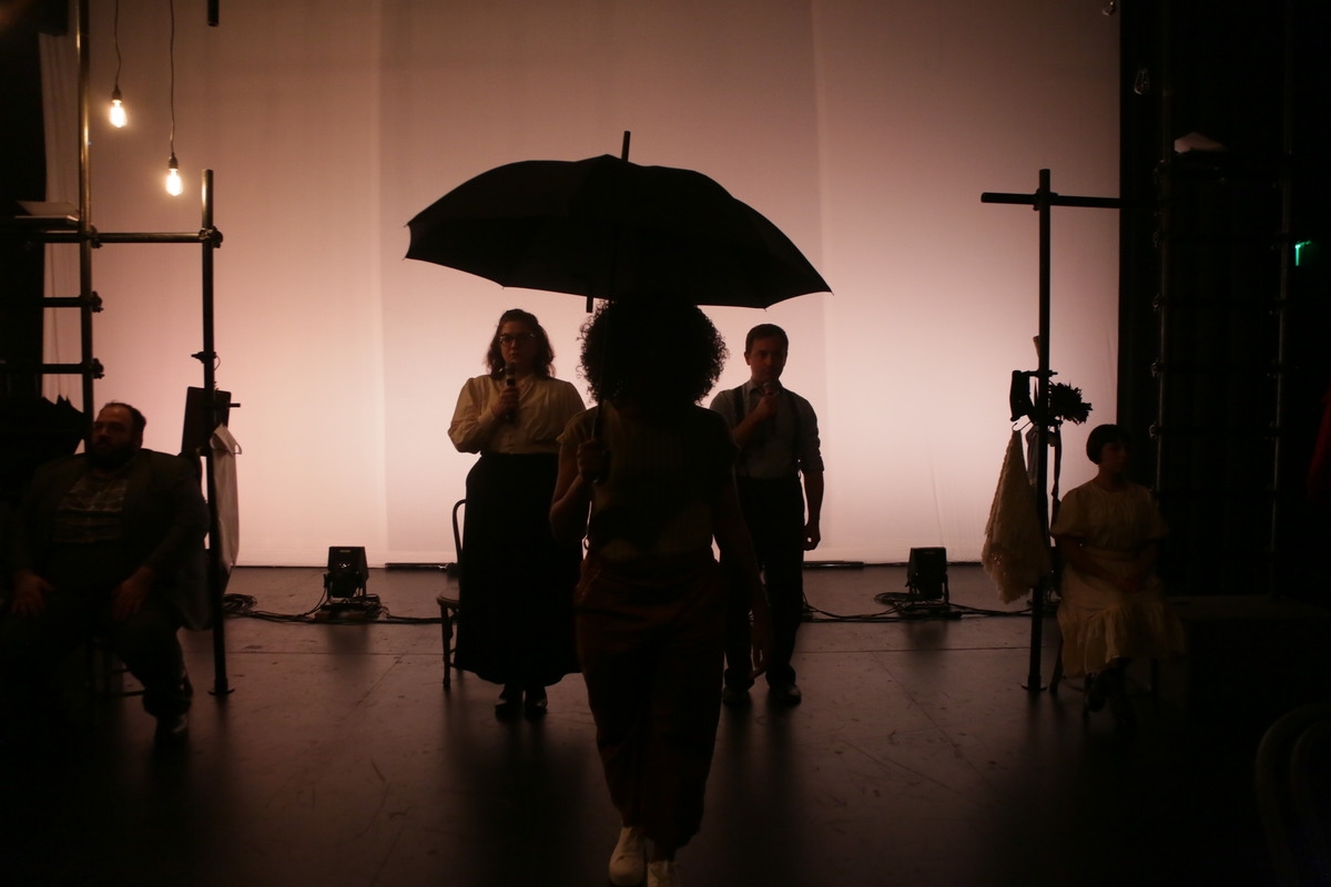Stage set, lit in dark pink, with metal utility carts at either side. Five people--one each sitting on a cart, two in middle, each holding mikes, and one in front holding an umbrella--populate the stage.