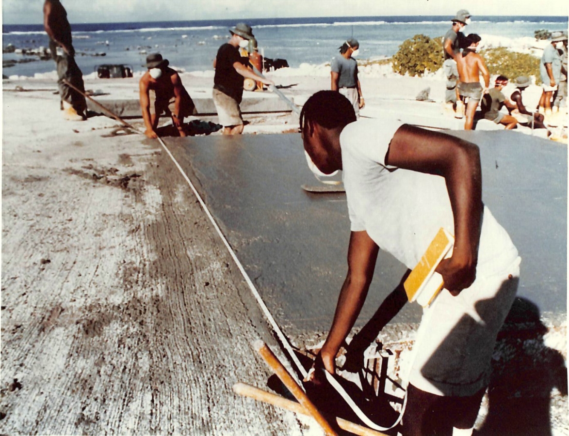Young Black man, seen from behind, stands on expanse of concrete and bends down while holding measuring tape. 