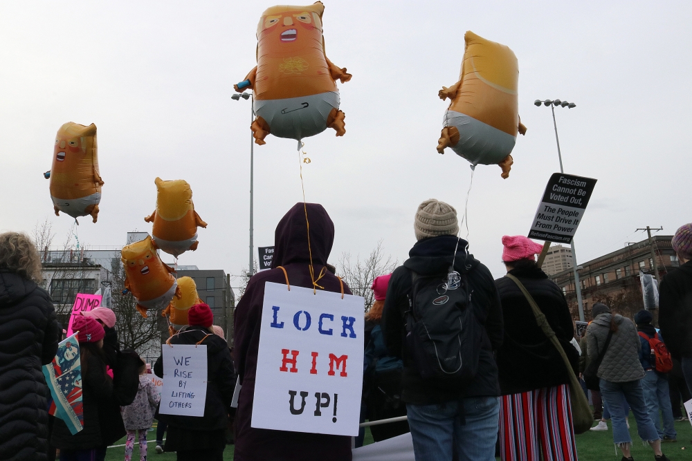 Shelley Mortinson, left, of Indivisible Marysville wears a “Lock Him Up” sign listens to rally speakers while flying her Trump baby balloon. Photo by Jon Williams