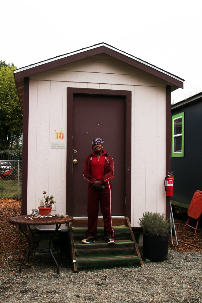 Young Black woman in track pants, jacket, and head kerchief stands on steps of a tiny house with planters on either side of path.