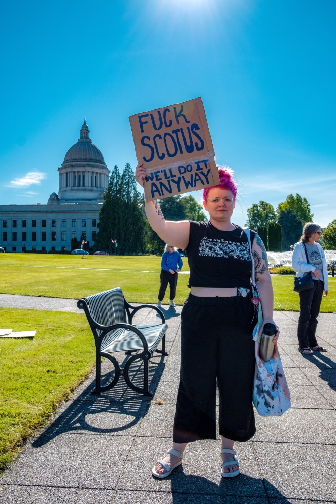 Young white woman wearing glasses, hair in topknot, and cut-off T-shirt with pro-choice message holds sign reading, "Fuck SCOTUS We'll do it anyway."