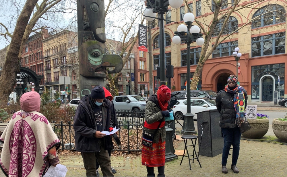 Four people in coats and hats stand in front of Native art sculpture on street in Pioneer Square.