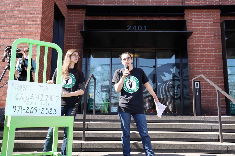 Young white man and woman stand on steps, man talking into a microphone, behind a green chair that bears a sign inviting people to call a number if they want to organize