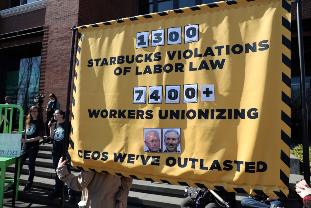 People hold banner reading, "1300 Starbucks Violations of Labor Law," "7400+ Workers Unionizing," and "CEOs We've Outlasted," under two photographs of older white men, Starbucks CEOs
