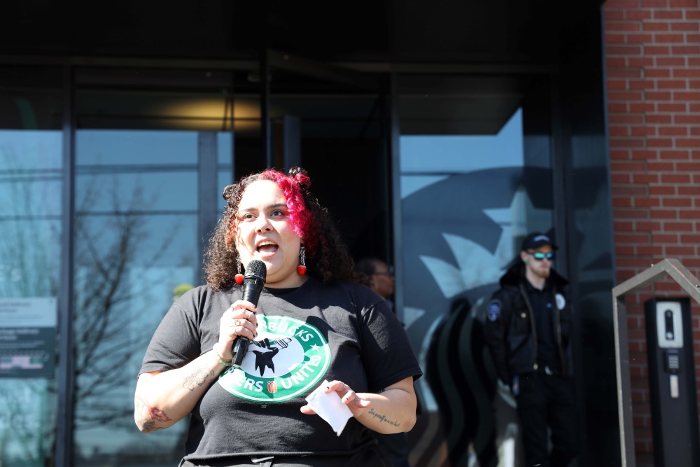 Young woman with curly dark hair dyed pink in front, wearing Starbucks Workers United T-shirt, talks into a microphone.