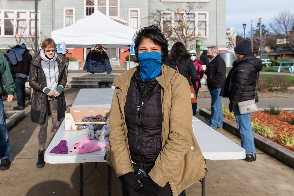 A woman with tan skin, short dark hair, wearing a blue neck gaiter and tan coat over black jacket, stands in front of table displaying hats, gloves, and other items for donation