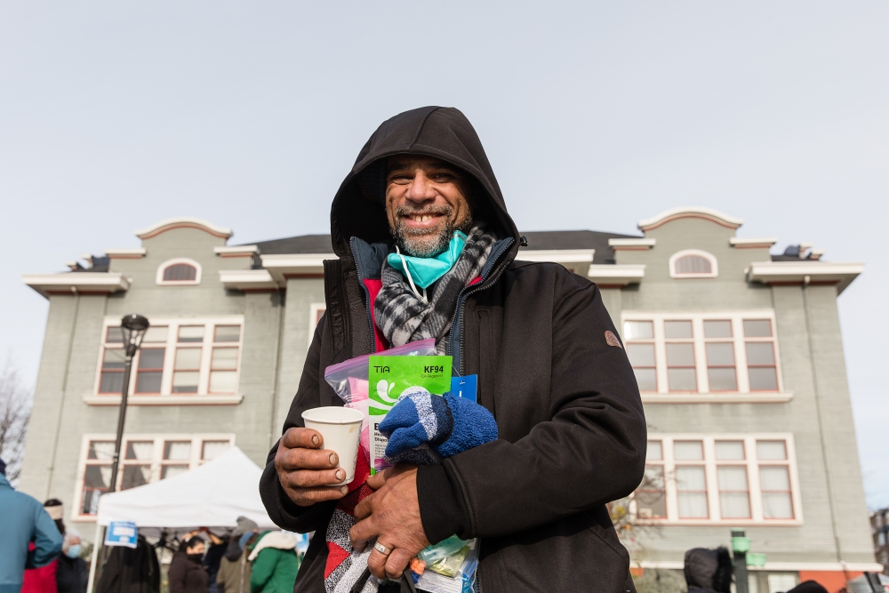 Smiling middle-aged, bearded white man in hooded black coat holds coffee cup in one hand and donated items in plastic bags in the other.