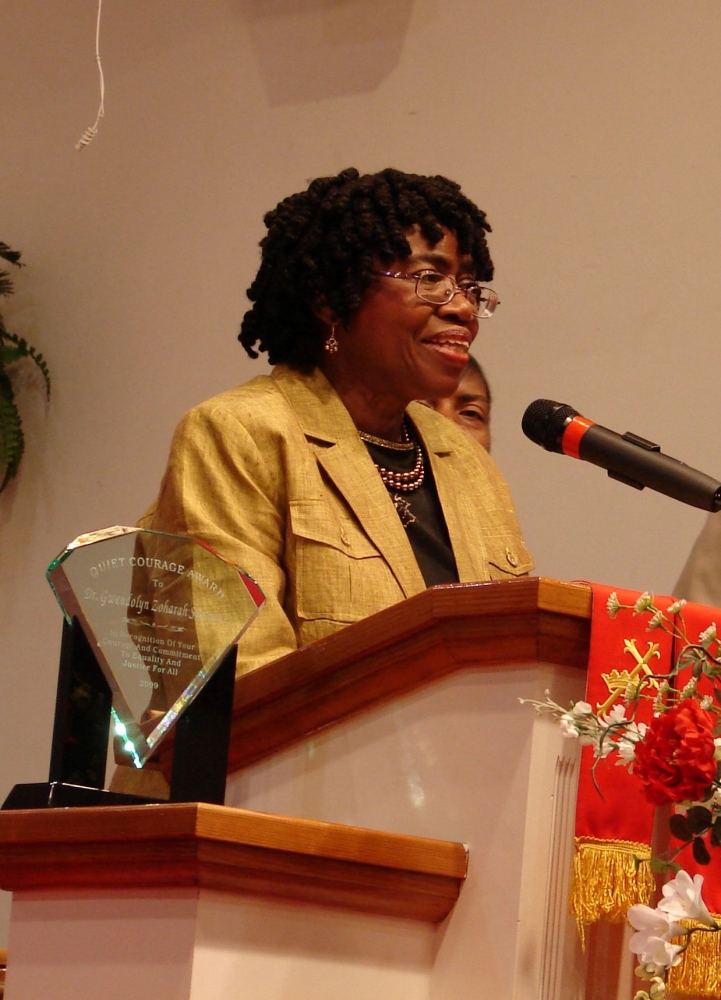 Middle-aged Black woman with glasses and short dreadlocks speaks at a podium.