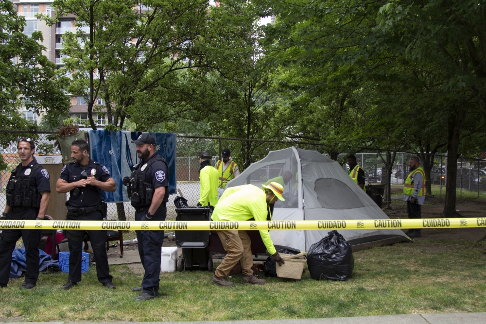 Four uniformed Seattle Police Department officers stand behind a strand of yellow caution tape on a dirt and grass lawn dotted with trees in Seattle's Denny Park, as city workers in yellow safety vests finish packing up a camper's tent and belongings.