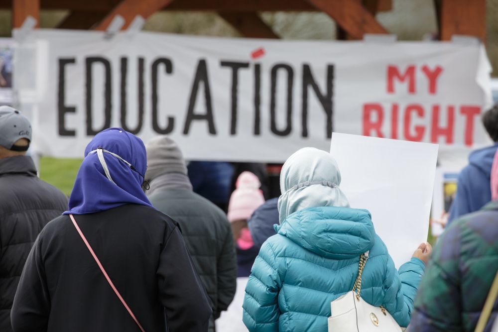 Rally attendees, seen from behind, in jackets and hijabs, in front of a banner that reads, "Education My Right"