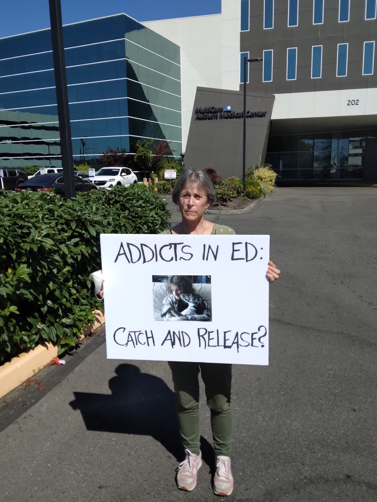 Woman stands next to hedge outside MultiCare facility, holding sign that reads, "Addicts in ED: Catch and Release?"