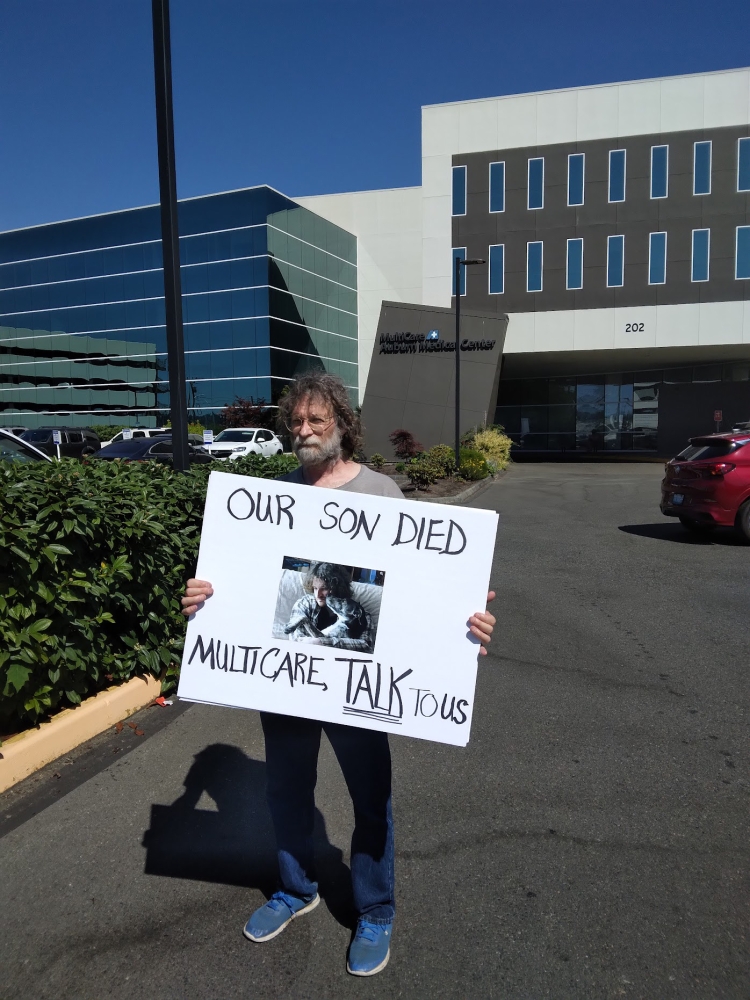 Man stands outside MultiCare facility holding sign that reads, "Our Son Died--MultiCare Talk to Us."