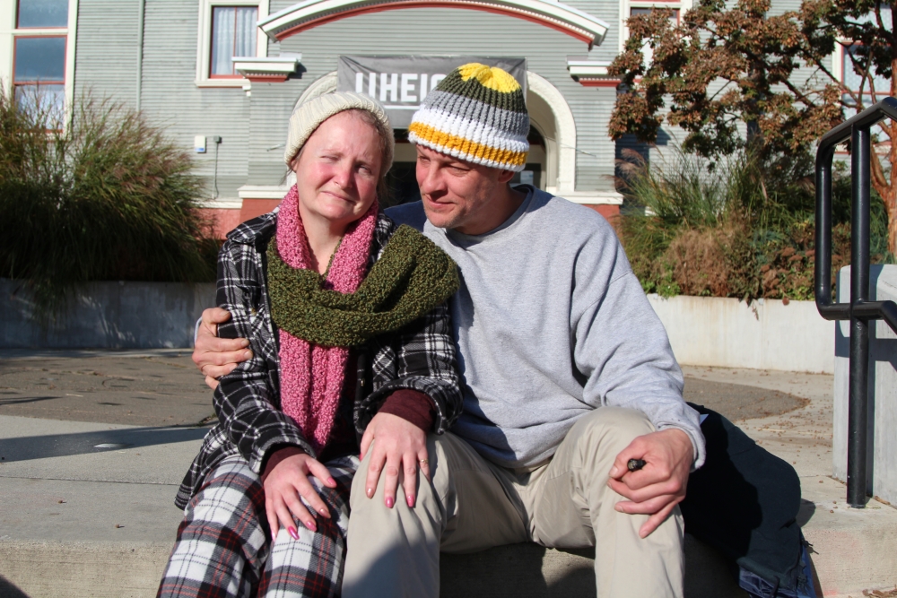 Middle-aged white couple, man holding arm around woman, wearing knit hats and thick shirts, sit in front of U Heights community center on sunny day