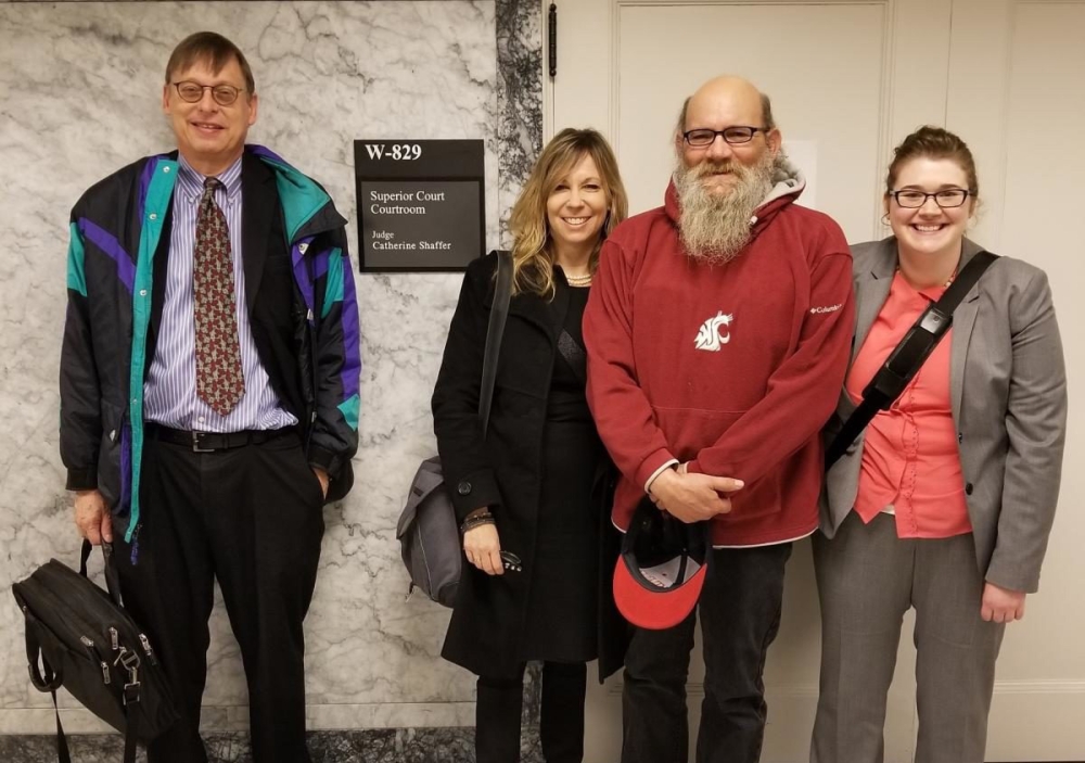 Two men and two women stand in a court office corridor. The man third from left wears glasses, a long gray beard, and a red hoodie showing WSU logo